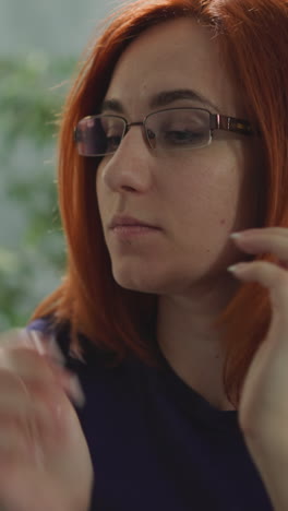 young woman with ginger hair puts on glasses to continue working on computer in office. female employee takes care of eyesight using eyewear closeup