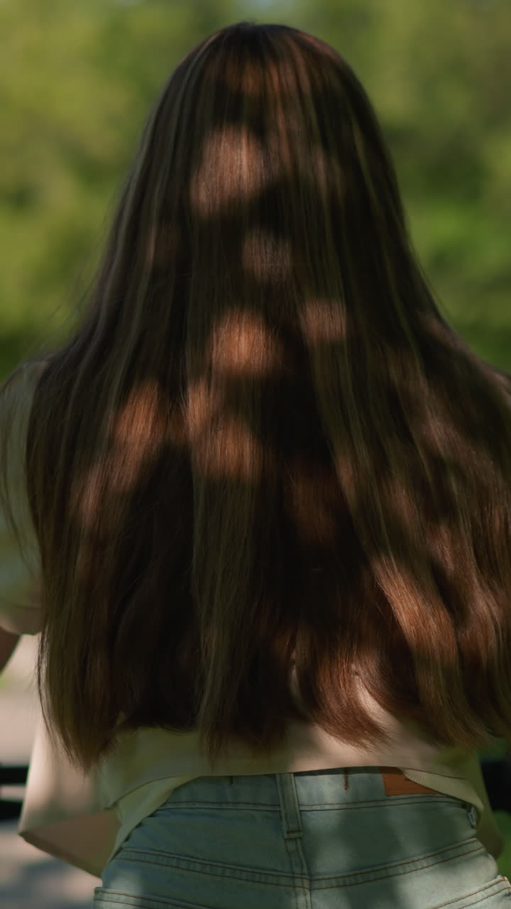 풍경적인 공원을 통해 자전거를 자신감 있게 타고 있는 젊은 여성의 뒷면, 긴 머리카락이 그녀의 등 아래로 떨어지고, 나무의 그림자가 그녀의 머리카락과 옷에 패턴을 만니다.