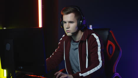 Close-Up-View-Of-Young-Serious-Man-With-Headphones-And-Playing-A-Game-On-The-Computer-In-A-Room-With-Colorful-Neon-Lamps-On-The-Wall