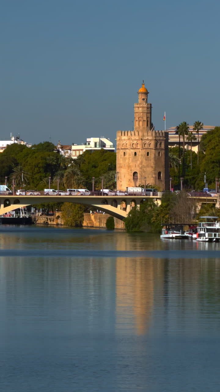 Ponte fluviale illuminato dal sole, riflesso di torre storica sotto un cielo azzurro limpido, specchio d'acqua calma con leggere increspature.