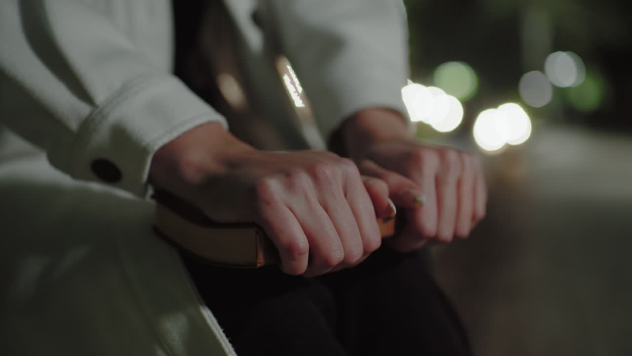 Close up lady in white coat firmly gripping book with bright bokeh lights in distant, capturing serene night moment, gentle nails pressed on cover, softly illuminated background
