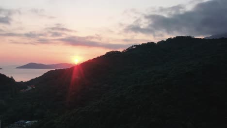 Aerial-truck-left-of-sunset-sunbeams-behind-rainforest-hills-and-village-in-Pui-O-bay,-sea-in-background,-Lantau-Island,-Hong-Kong,-China