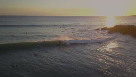 Vista-Aérea-De-Un-Surfista-Monta-Las-Olas-Del-Océano-En-Longboard-Con-Velocidad,-Atacando-El-Borde-De-La-Ola-Y-Generando-Una-Impresionante-Cantidad-De-Rocío-Con-Una-Enorme-Puesta-De-Sol-En-El-Fondo