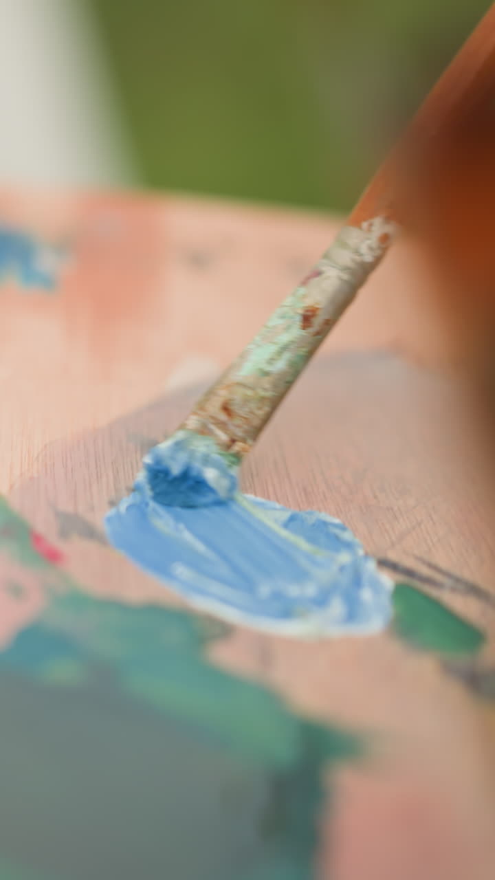 An artist s hand carefully mixes blue and white paint on a wooden palette using a brush. The close-up shot highlights the delicate process of blending colors, with various other hues visible