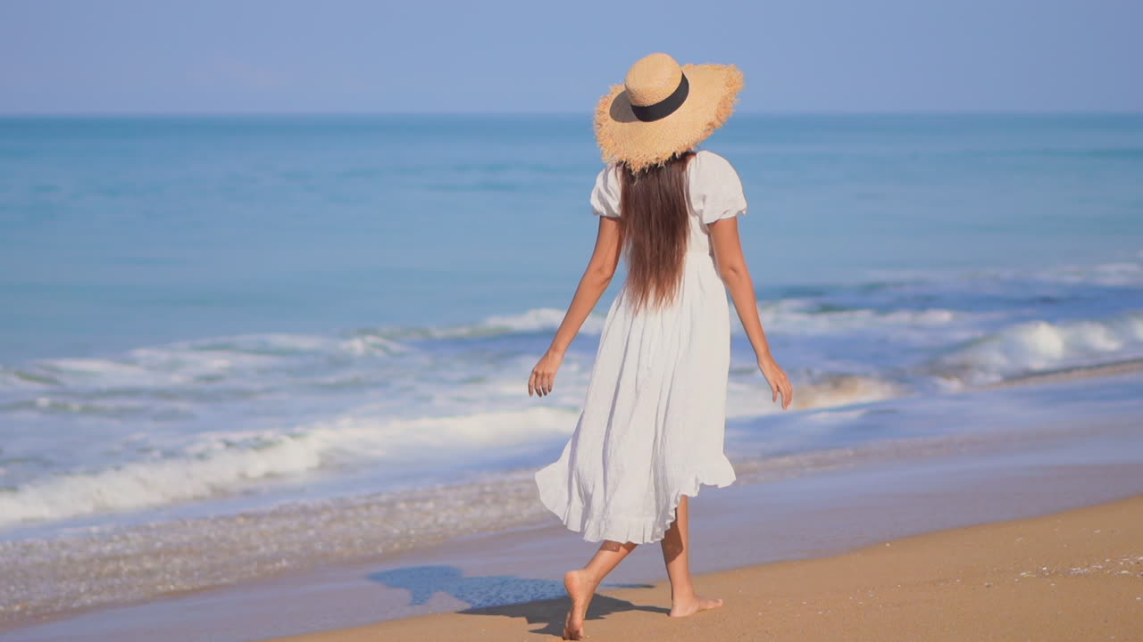 선드레스와 플로피 밀짚 모자를 쓴 사랑스러운 젊은 여성이 파도가 밀려올 때 해변을 따라 걷는다