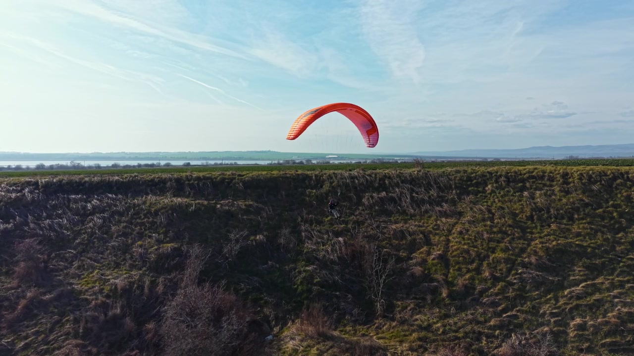 Paraglider soaring over a grassy hill near the water on a clear day with few clouds in the sky