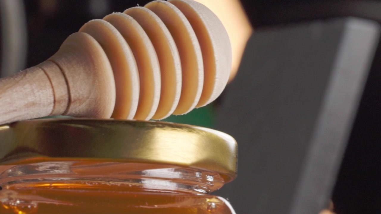 Primo piano panoramico di un mestolo di legno per il miele appoggiato su un barattolo di vetro pieno di miele dorato, che evidenzia la dolcezza naturale e i prodotti dell'apicoltura