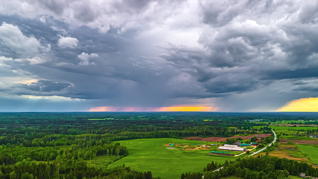 Dark rain streaks down and light dance across hills and fields under shifting cloud cover with orange golden sky behind light rays
