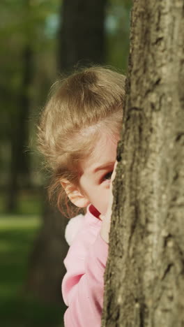 playful little child hides behind old trunk in spring garden closeup slow motion. pretty blonde girl has fun peeking out from behind tree. happy time in sunny park