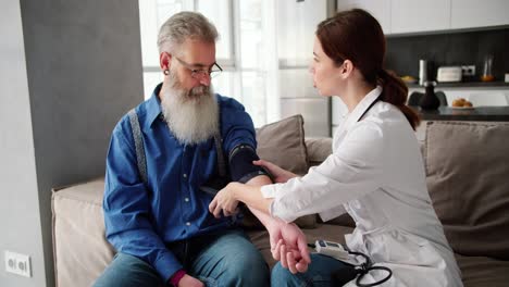 a brunette girl in a white coat the doctor expresses her verdict after an examination with a heart rate monitor and a blood pressure examination of an elderly man with gray hair and a lush beard in a blue shirt on the sofa at home