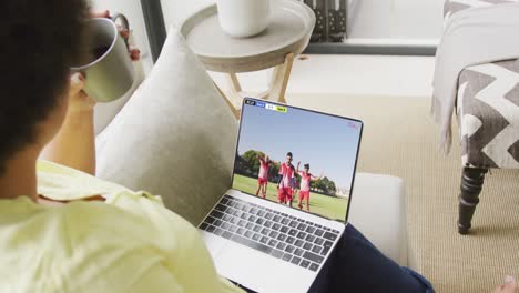 video de una mujer biracial sentada en el sofá y viendo fútbol en la computadora portátil en casa