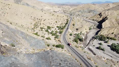 Aerial-drone-zoom-in-shot-of-vehicle-driving-along-winding-RCD-Road-surrounded-by-arid-vegetation-in-Balochistan-on-a-sunny-day