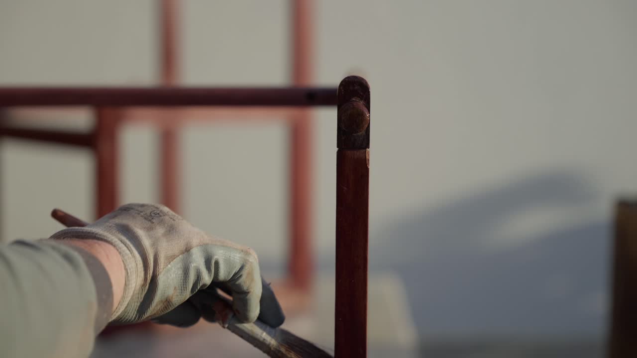 Close up of gloved hand using brush to apply varnish on vertical wooden chair leg outdoors in daylight