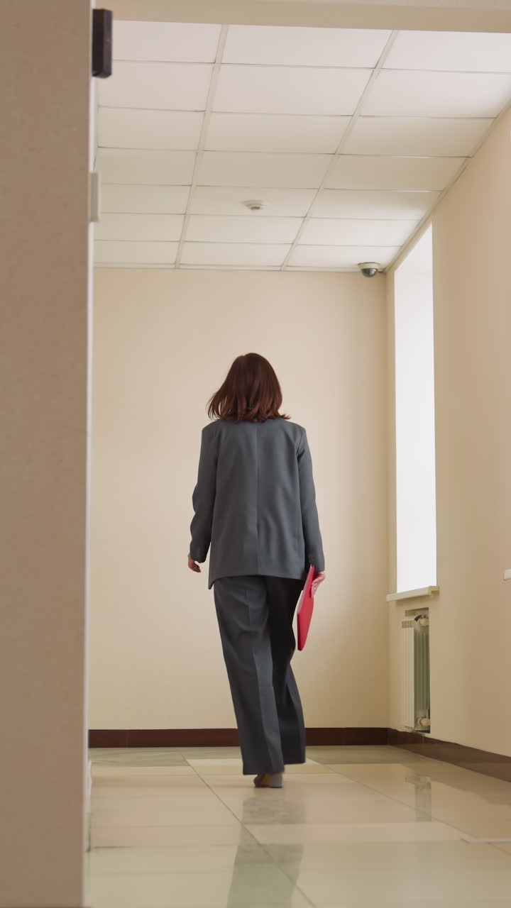 Rear view of confident businesswoman walking alone down quiet office corridor in formal suit toward meeting room, suggesting ambition, purpose, career progression, professional mindset