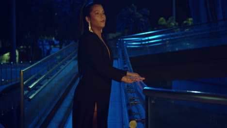 a woman in a black dress leans on the railing of a walkway in the city at night