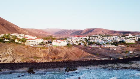 ciudad costera enclavada contra las colinas bajo cielos despejados, olas golpeando costas de arena oscura, luz serena de la noche, vista aérea