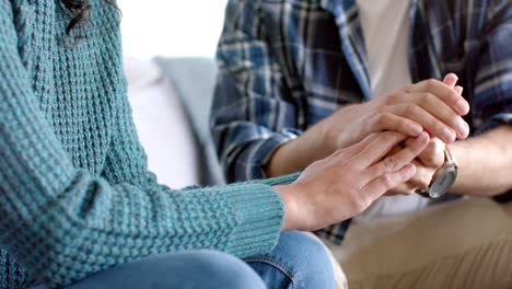 Midsection-of-biracial-couple-sitting-on-sofa-and-holding-hands-at-home,-in-slow-motion