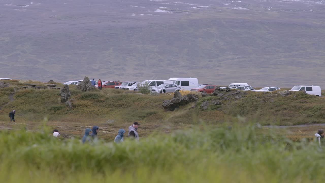 Tourists walk through a scenic landscape in Iceland with parked cars in the background