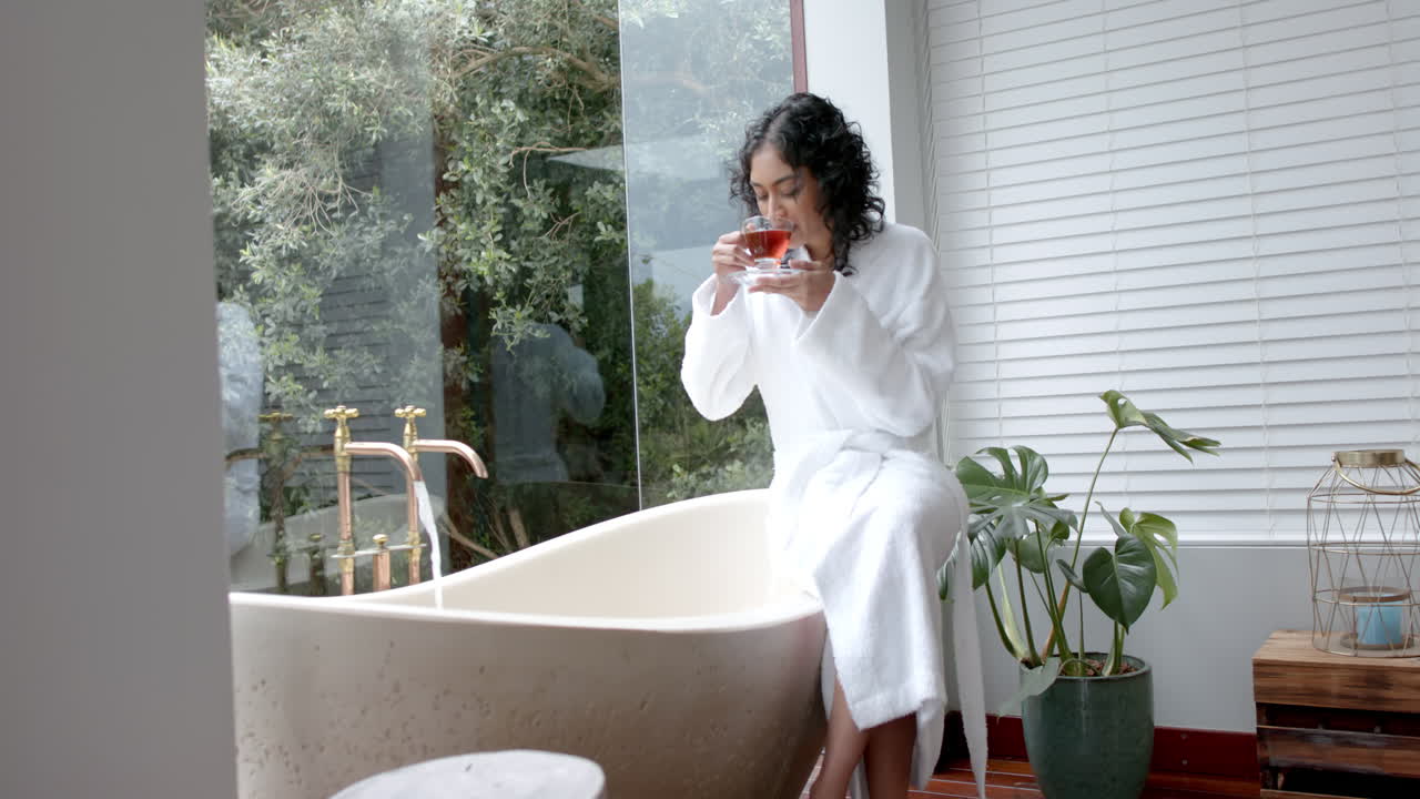 двухрасовая женщина в халате сидит на ванной, пьет чай в ванной в доме, замедленная съемка