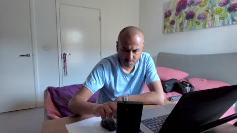 South asian man concentrating on his remote work, using laptop and checking smartphone in his bedroom
