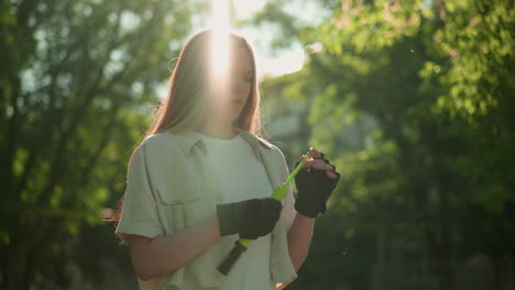 lady wearing black gloves stands outdoors, thoughtfully observing nozzle of green pump in hand, surrounded by a background of trees and a distant building