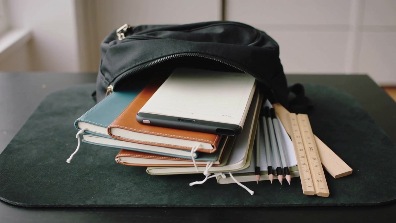 Unzipping black pouch sending notebooks, slim device, ruler and pencils onto green mat at home desk
