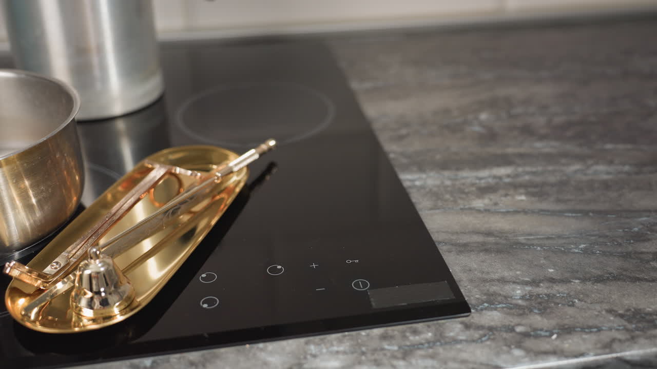 Gas cooker with stainless pot and golden baking tray on black glass surface as kitchen towel is dropped beside tray, capturing moment of food preparation and cleanup in modern cozy kitchen setup