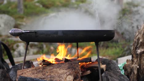 close up shot of muurikka traditional finish classic griddle pan, over a hot wood fire and steaming water being poured over, in a camping environment