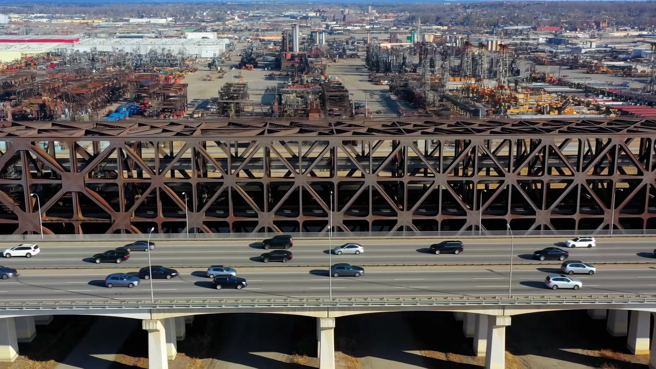 Moving cars pass across large steel bridge roadway while distant buildings fill horizon, capturing urban transportation, structural details, and open daylight view from elevated perspective.