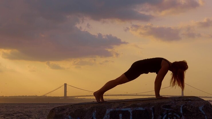 Donna che esegue la posizione yoga del cane a testa in giù con lo sfondo di un ponte di New York. Silhouette laterale di una donna che si allunga su un molo sul lungomare di fronte al punto di riferimento Verrazzano Narrows al tramonto.