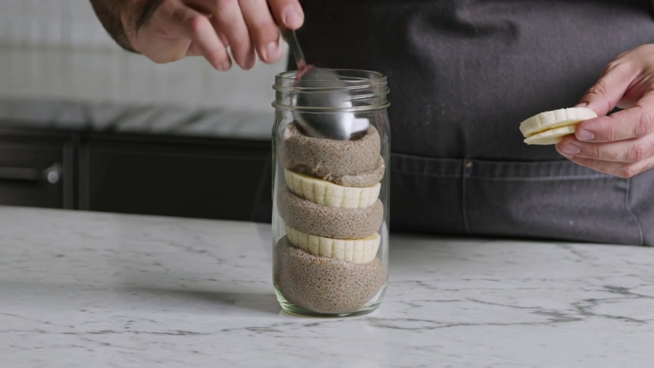 Un cuoco con un grembiule scuro riempie un barattolo di vetro con un cucchiaio su un piano di marmo, preparando uno spuntino con fette di banana.
