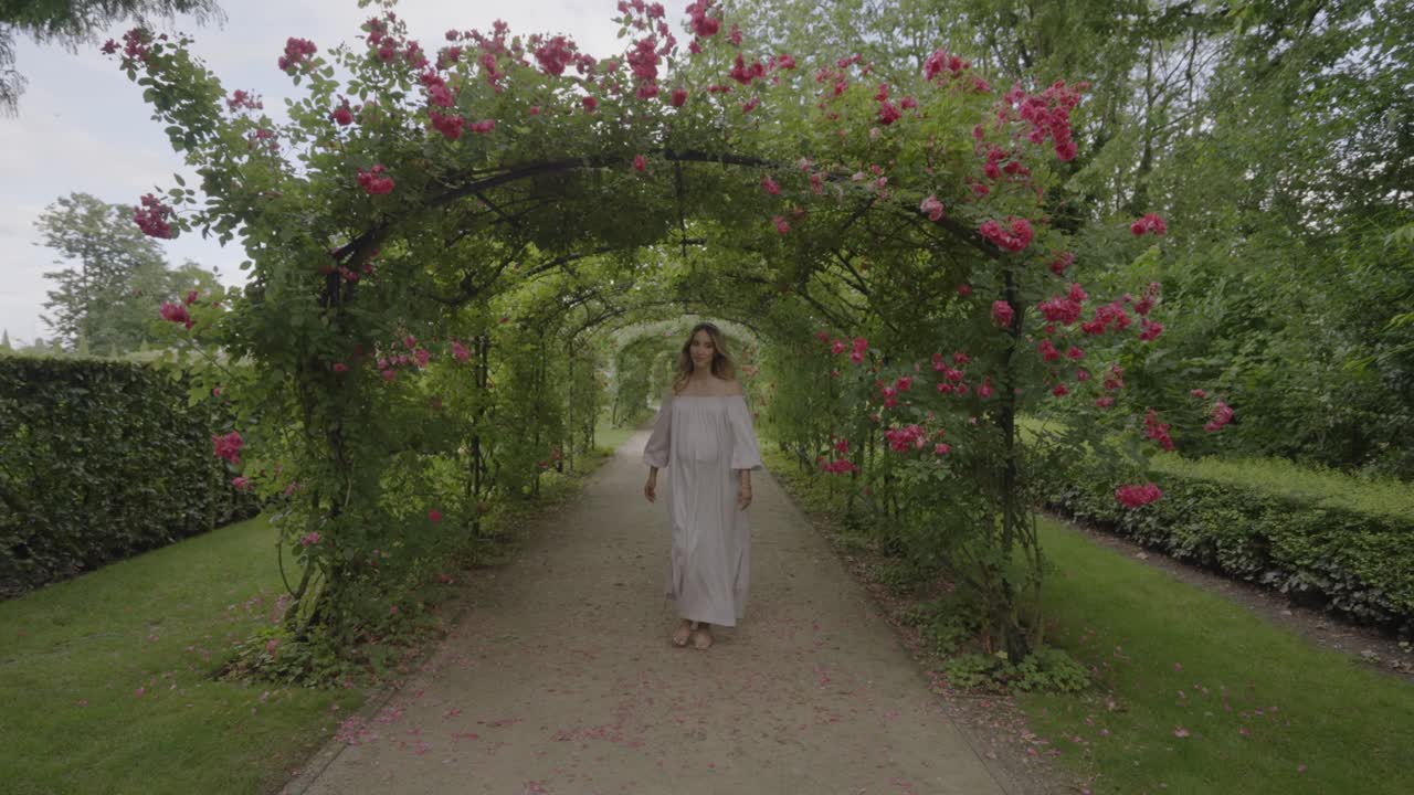 Elegant young pregnant lady in a beautiful white dress walking through a rose tunnel, dancing happily and rubbing her belly, 4K