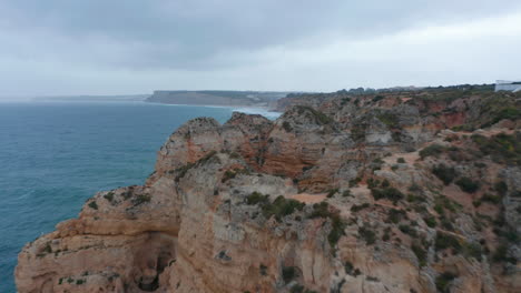 Aerial-drone-flying-forward-near-rocky-coloured-cliffs-in-Lagos,-Portugal,-along-coast-with-lighthouse-on-background,-cloudy-day