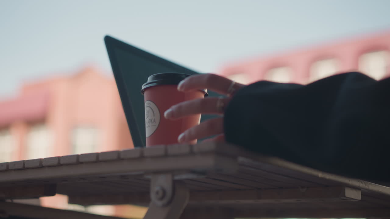 손가락에 반지를 달고 커피 컵을 들고 있는 사람의 클로즈업, 테이블 위에 노트북 에 놓고, 노트북 키보드와 화면이 보이고, 건물의 흐릿한 시야와 밝은 하늘을 배경으로