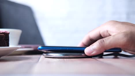 persona cargando un teléfono inteligente azul en un cargador inalámbrico en una mesa de madera con comida y portátil en el fondo