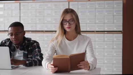 blonde woman reads book near black man using laptop. lady takes off glasses to have break during learning material for future exams. student hard work