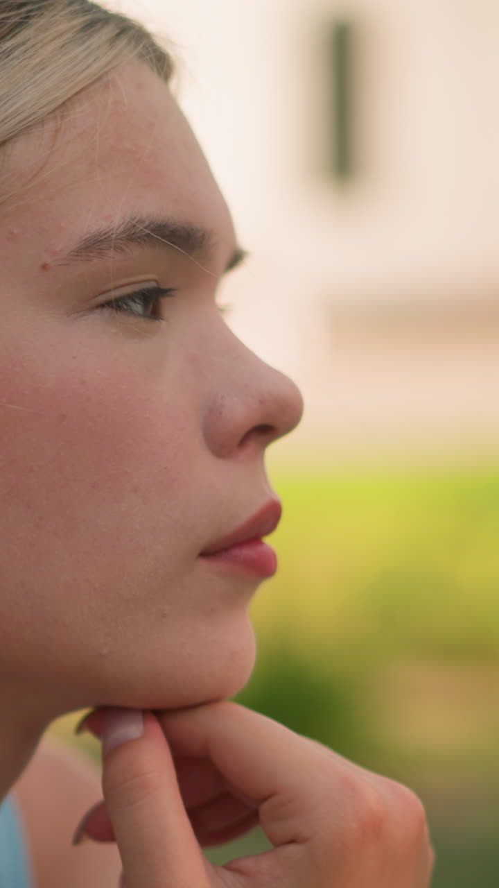 vista lateral de una mujer joven con la mano debajo de la barbilla, mirando pensativa mientras mira hacia la distancia, con fondo de vegetación borrosa y edificio
