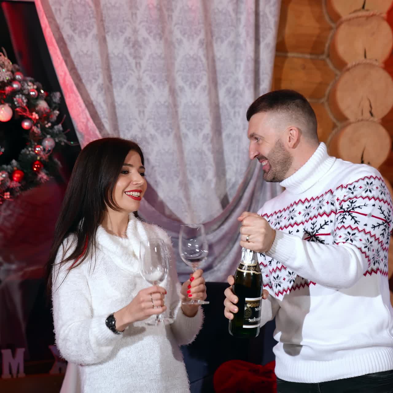Husband and wife celebrate New Year. Man and woman in white sweaters hold glasses and champagne in Christmas decorated room. Romantic atmosphere