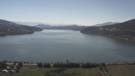 Luftüberflug-über-Autoschleppjacht-Durch-Ein-Bergtal-Am-Seeufer-Mit-Einem-Smogigen,-Nebligen-Horizont-Mit-Glitzerndem-Wasser-Und-Perfekt-Gepflegten-Gartenhäusern-An-Einem-Sonnigen-Sommernachmittag