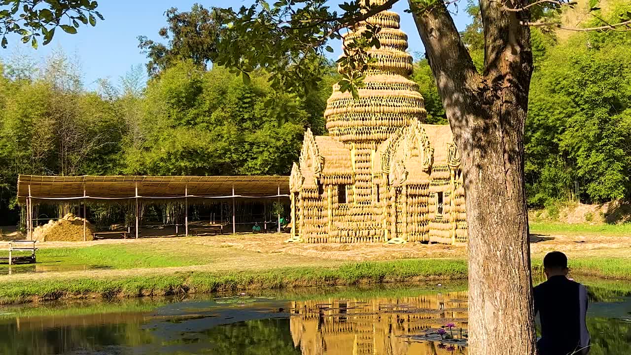 A rustic straw building reflected in a calm pond, surrounded by lush greenery and a single tree.