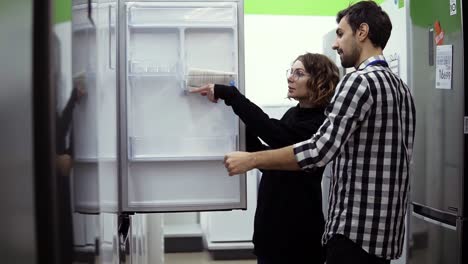 young married couple inspect open door refrigerator, design and quality before buying in a consumer electronics store