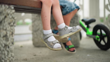 a close-up of two young children's legs as they casually swing their feet back and forth, one leg is crossed, and a green bicycle is parked nearby