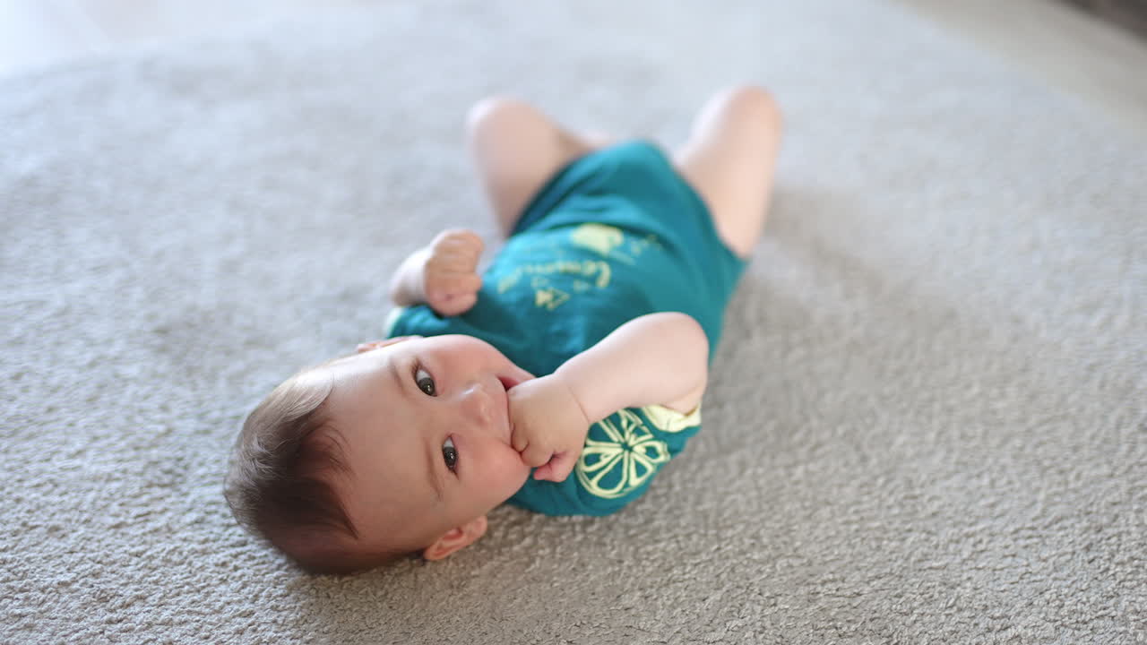 Lovely kid lies on the floor his head to the camera. Baby chewing his fist and smiling sweetly. Blurred backdrop.