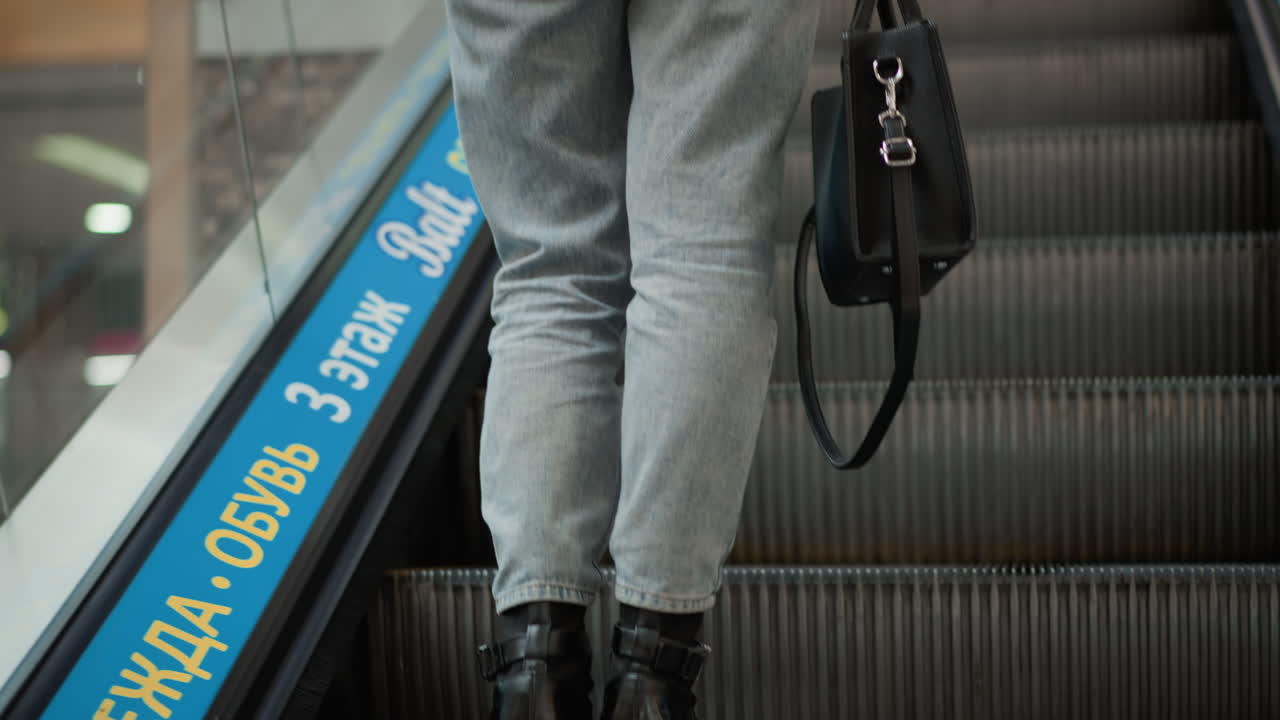 back view of female shopper stepping onto moving walkway in modern mall hall wearing white sweater under bright lights next to glass railing capturing dynamic motion