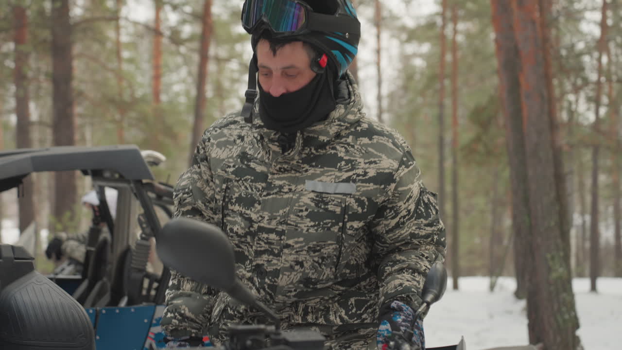 Caucasian man prepares atv snowy forest expedition guide adjusts helmet and goggles, buckles balaclava, inspects blue utv beside pine trees, visible breath in cold, focused calm before ride