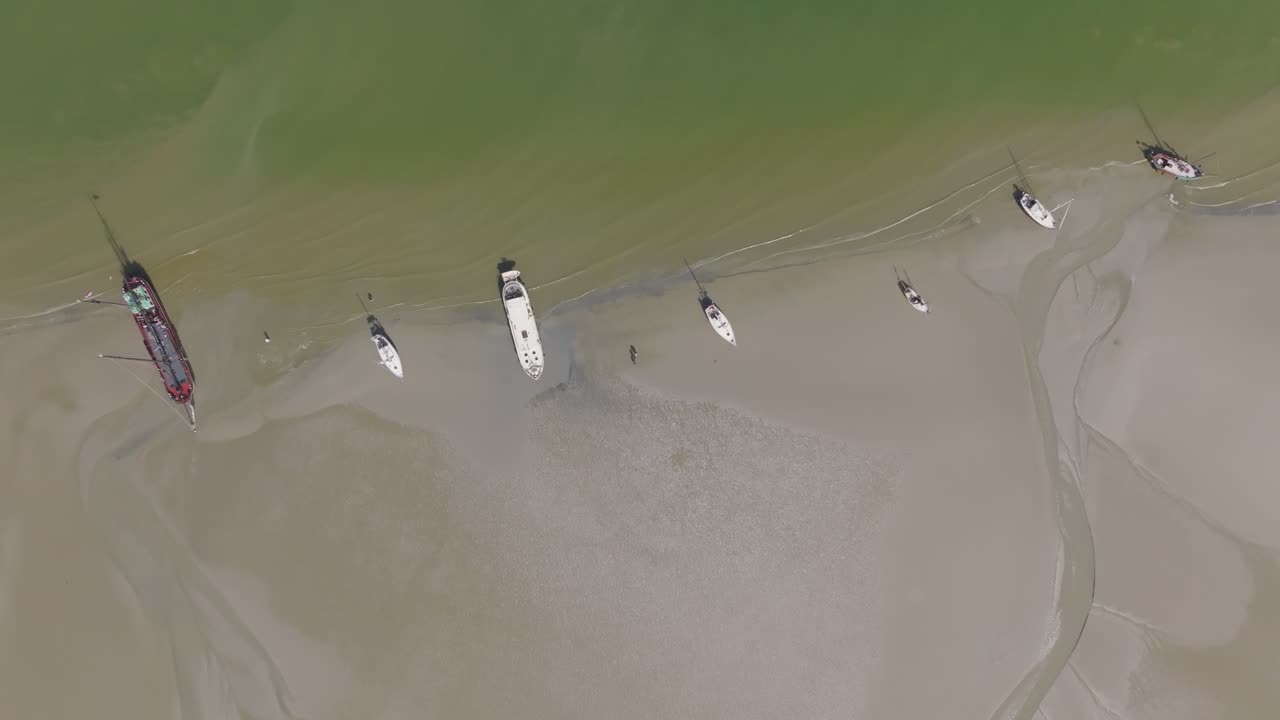 Overhead view of yachts, a classic barge and catamaran aligned along green water and sand channels; drone holds a true vertical composition. Wadden Sea