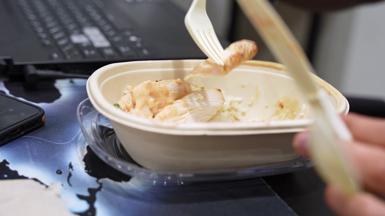 Una persona che mangia una ciotola di riso al pollo con salsa a una scrivania di lavoro. Un laptop e uno smartphone sono visibili sullo sfondo, raffigurando una pausa pranzo d'ufficio impegnata o uno stile di vita da lavoro remoto