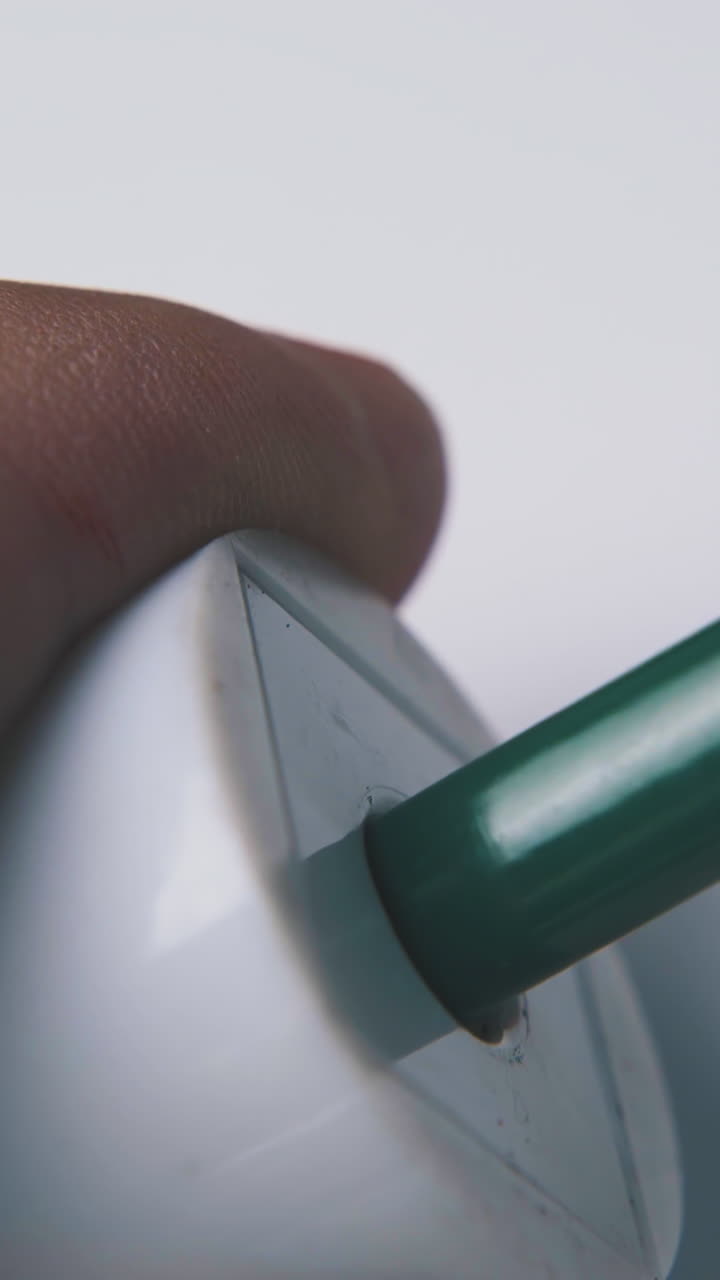 young man sharpens green color pencil with white plastic sharpener on light background extreme close view