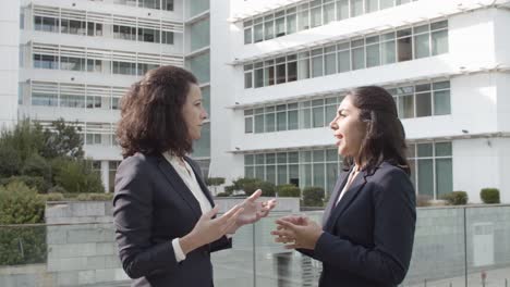 side view of two positive business colleagues standing outside office building, talking and gesturing, discussing work projects