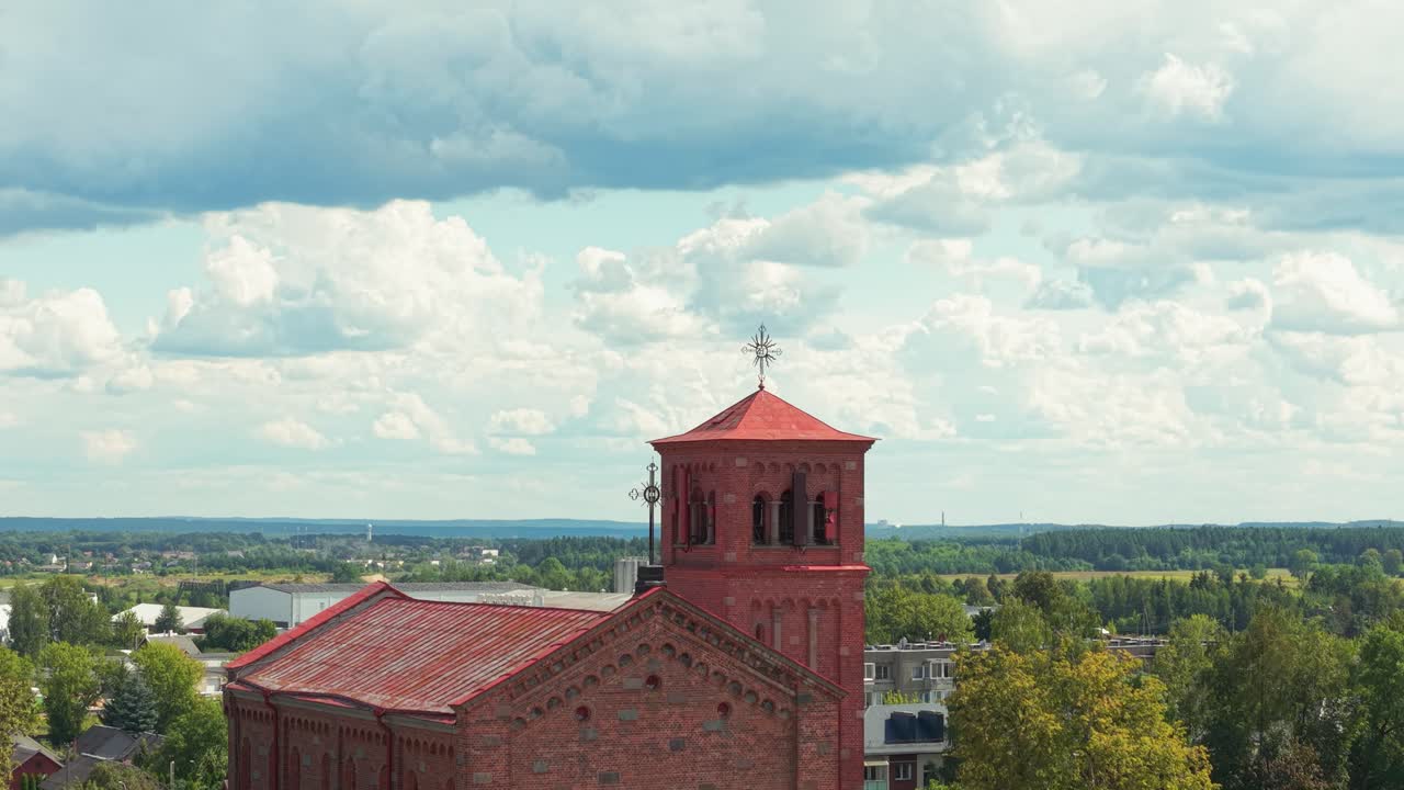 Aerial view of Lentvaris Church in Lithuania with its red brick architecture and bell tower, surrounded by lush greenery and a distant cityscape under a partly cloudy sky